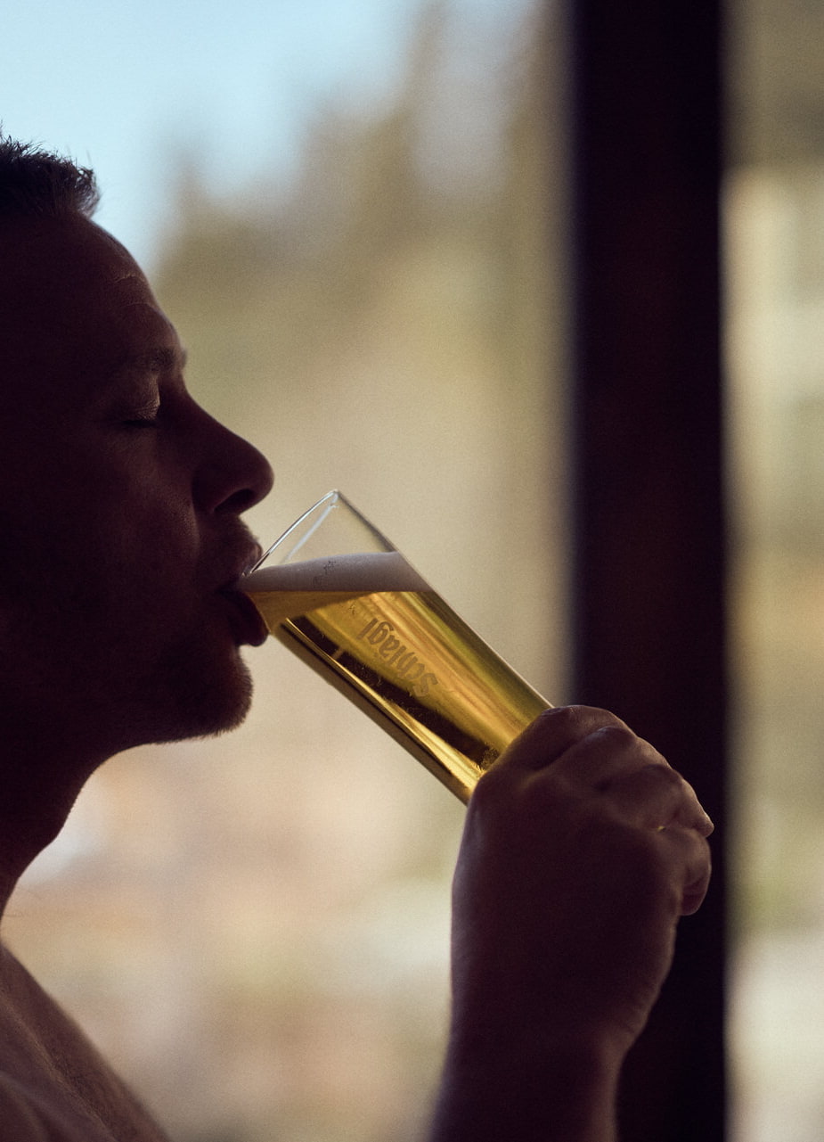 Ein Mann sitzt in der Sauna und trinkt ein Glas Bier. Im Hintergrund sieht man unscharf das Hotel Guglwald. 